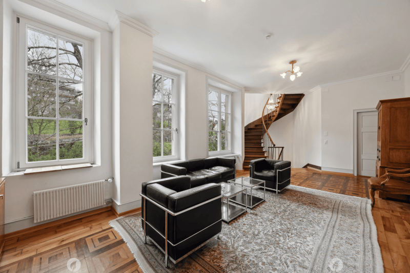 Open layout with two black leather sofas and glass tables, wooden floor, white walls, windows, staircase, chandeliers, radiator