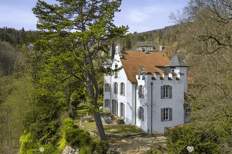 White house with brown roof, set on a hillside with multiple windows, small garden with fountain, surrounded by greenery