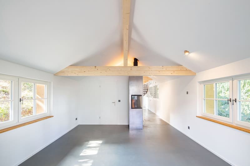 empty room with grey floor, white walls, wooden beams, fireplace, windows with wooden sills