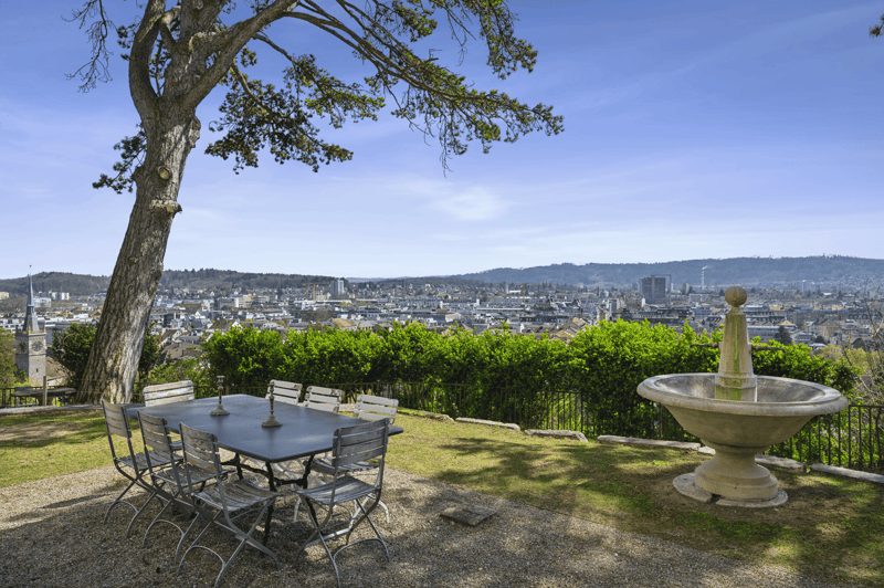 Outdoor dining table with chairs, small fountain, expansive city view, greenery