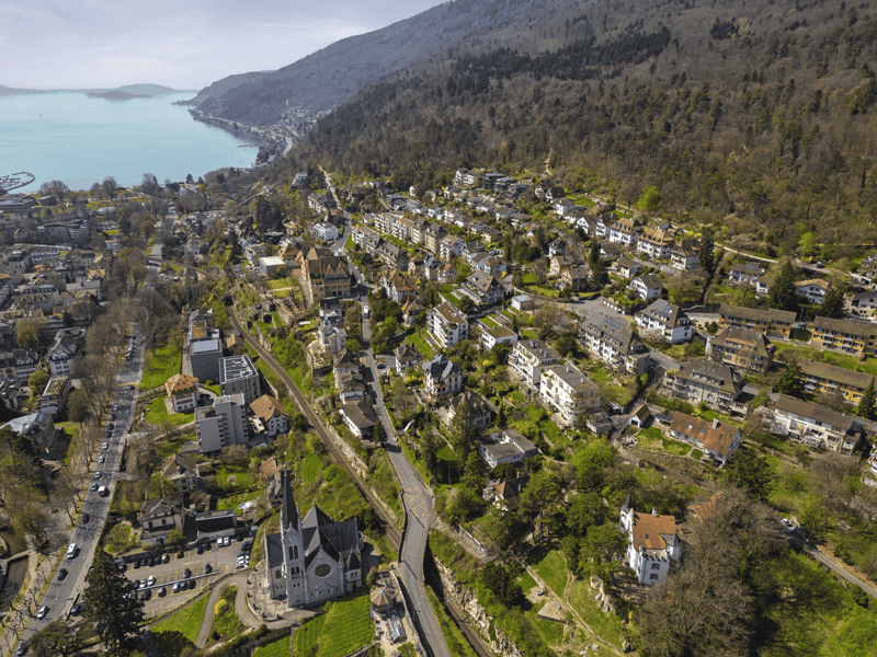 Panoramic aerial view of a hillside village with many houses, a church, and a winding road, with a body of water and mountains in the background.