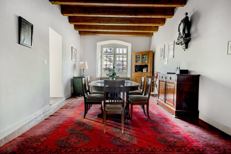 Dining room with large round table, wooden ceiling, white walls, wooden cabinets, floor lamp, large red carpet, windows