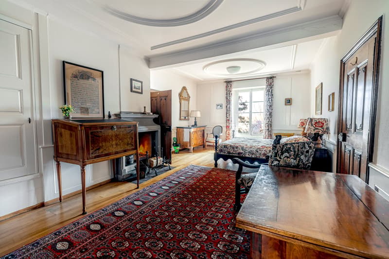 a wooden rug, a fireplace, a bed with floral bedding, a wooden desk, a chair, and a wall-mounted mirror