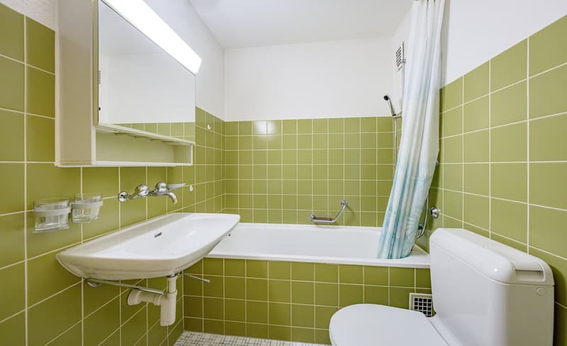 Bathroom with green tiles, white sink, bathtub, toilet, shower curtain, mirror, and glass holders on the wall.