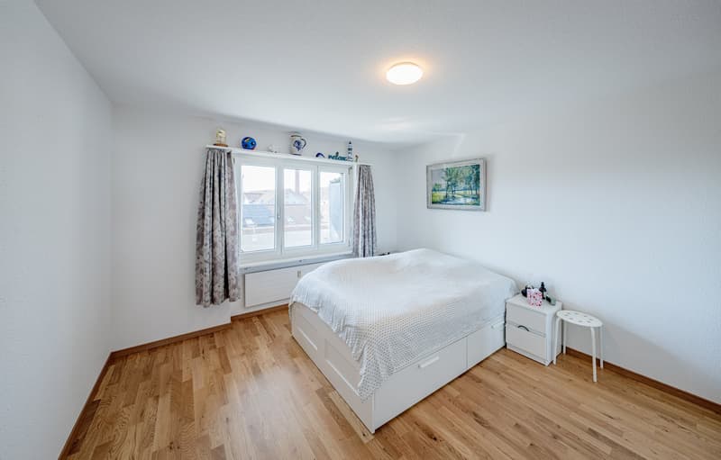 Bedroom with white walls, wooden floor, bed, nightstands, window with curtains, picture frame, ceiling lamp, and items on the window sill.