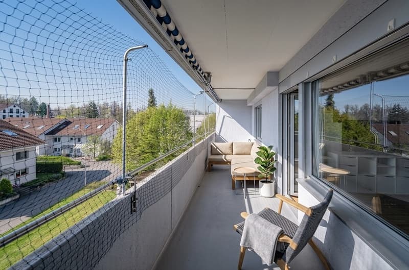 Balcony with a couch, table, chair, and potted plant, with a view of the street and houses
