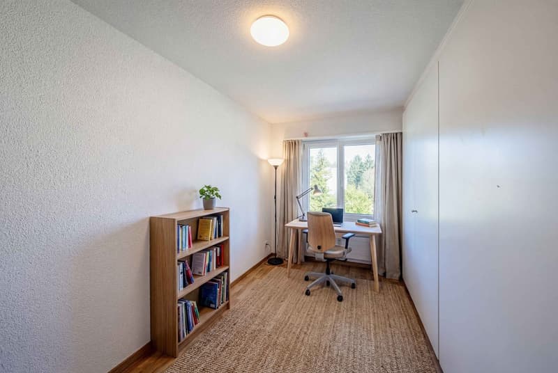 A room with a desk, chair, books, and a plant. The room has white walls, a wooden floor, and a window with curtains. There is a lamp on the floor and a ceiling light.