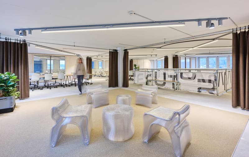 large open space, modern desks, chairs, suspended lights, white walls, beige carpet, decorative plants, glass partition with metal structure