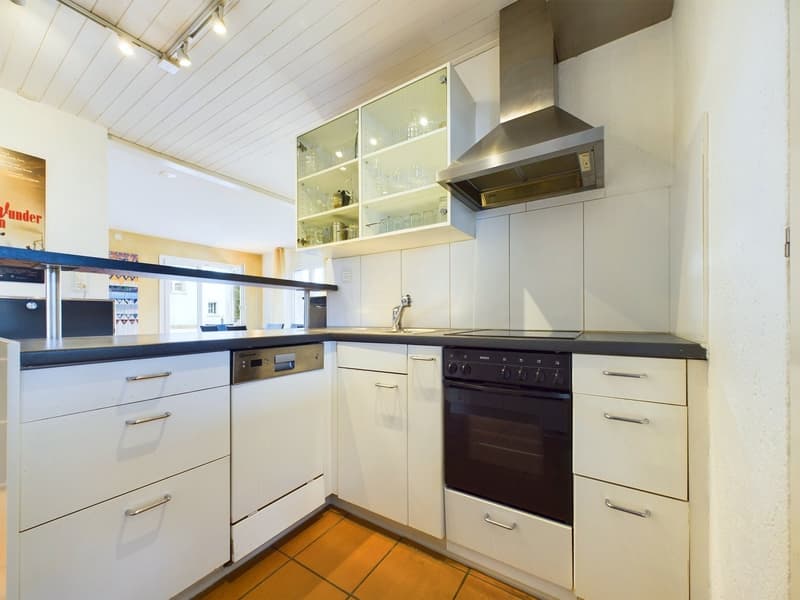 Modern kitchen with white cabinets, black countertops, stainless steel appliances, glass cabinets, tiled floor, overhead lighting, and a range hood.