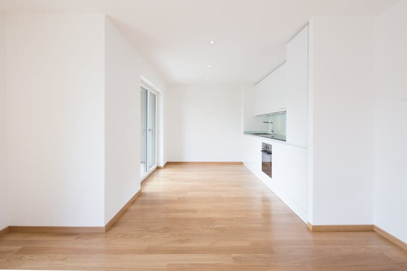 Empty room with wooden floor, white walls, large sliding glass doors, open kitchen, sink, oven.