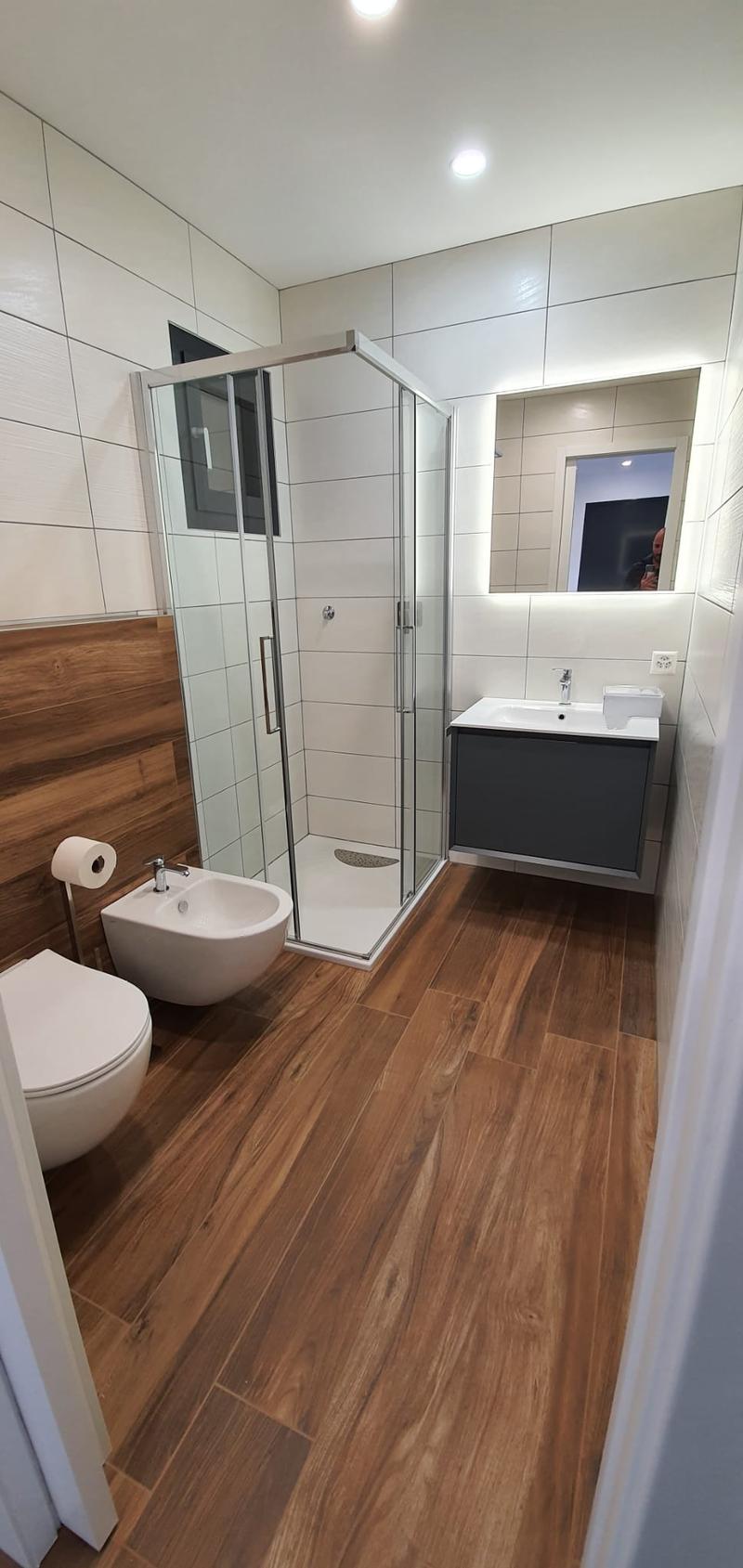 modern bathroom with wooden floor, white walls, glass shower, white toilet, white bidet, white sink, mirror