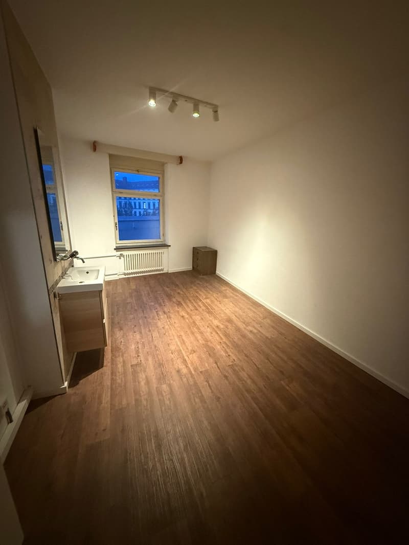 Empty room with wooden floors, large window, sink, radiator and storage box.