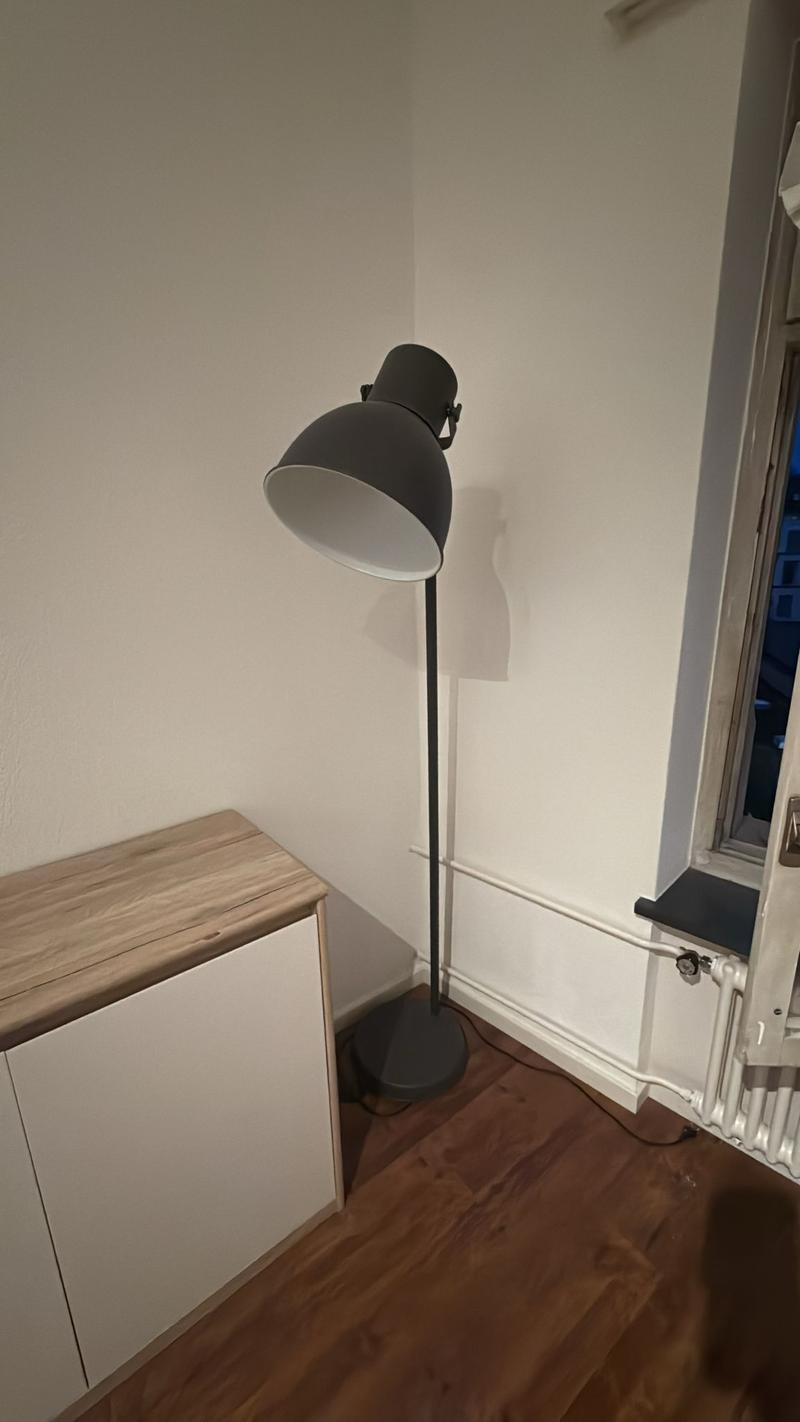 A white wall with a wooden floor, a tall grey lamp, a wooden cabinet, a window, and a radiator