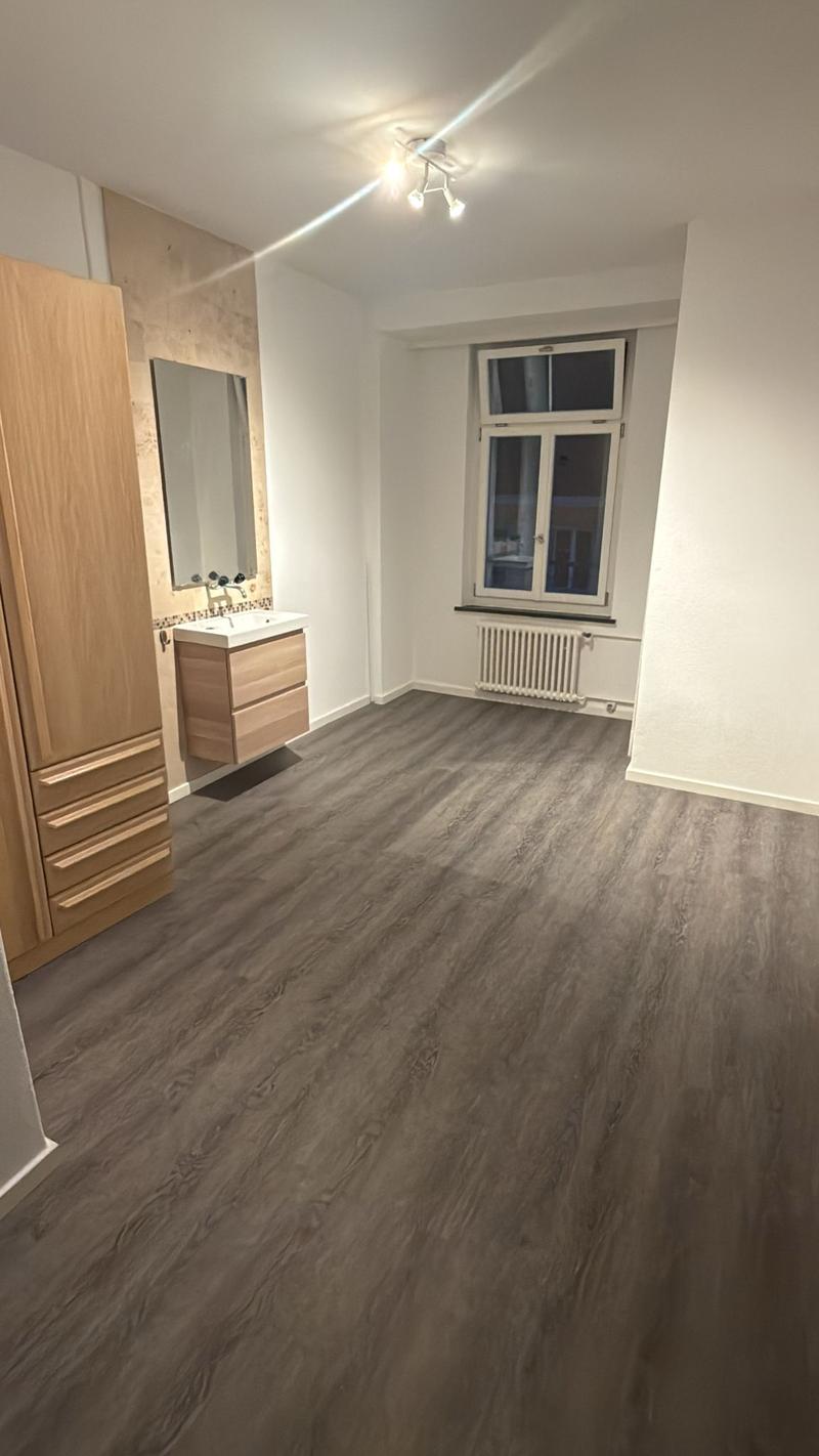 Empty room with hardwood flooring, white walls, wooden cabinet, mirror, sink, window with balcony, radiator