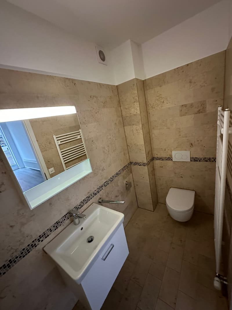 white tile walls, modern sink with faucet, white toilet, illuminated mirror, towel rack, tiled floor