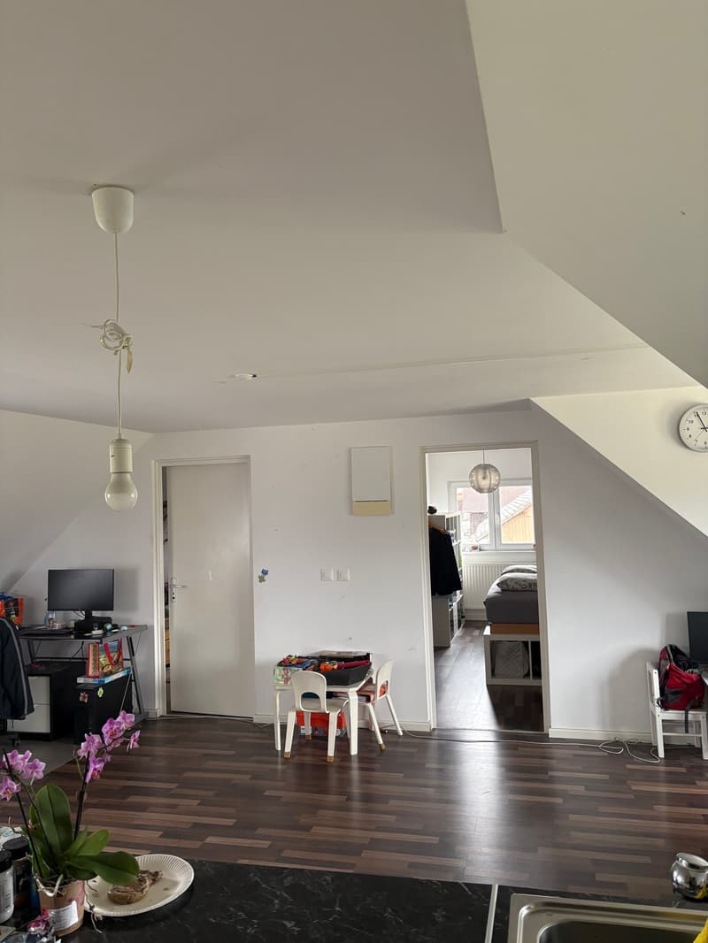 Living area with wooden floor, white walls, desk with monitor and flower, table and chairs, bed in the adjacent room