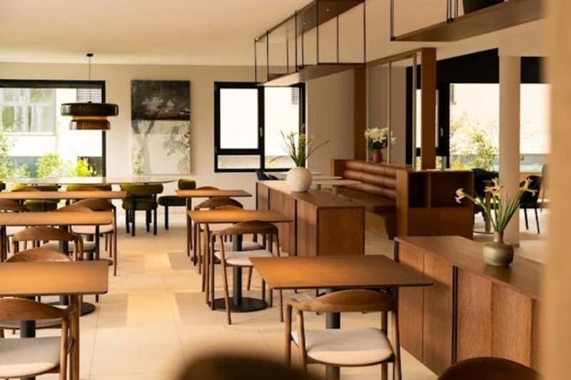 A cafe with tables, chairs, wooden benches, and a counter. There are potted plants, flowers, and a painting on the wall. The windows are large, allowing natural light to enter the space.