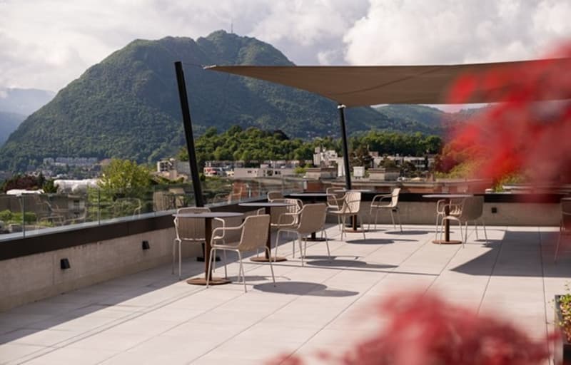 Outdoor seating area with tables, chairs, umbrella, plants, glass railing, mountain view