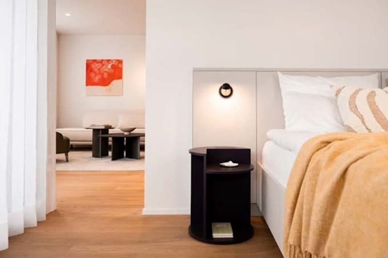 white walls, wooden floor, wall lamp, double bed with white pillows, dark nightstand, artwork, curtain