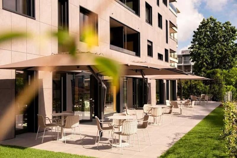 Outdoor dining area with tables and chairs, large umbrellas, paved area, green grass, buildings, trees