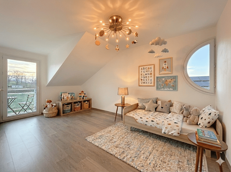 A cozy children's room with wooden floor, white walls, a round window, a daybed with pillows, a rug, a lamp, a shelf with toys, and a glass door leading to a balcony with a table and chairs.