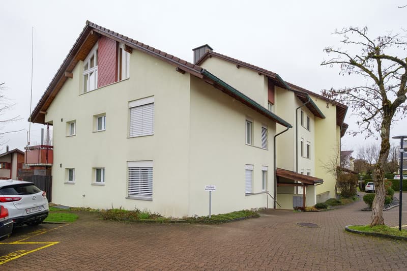 Apartment building, 3 story, white walls, red trims, window shutters, brown roof, cars parked in front