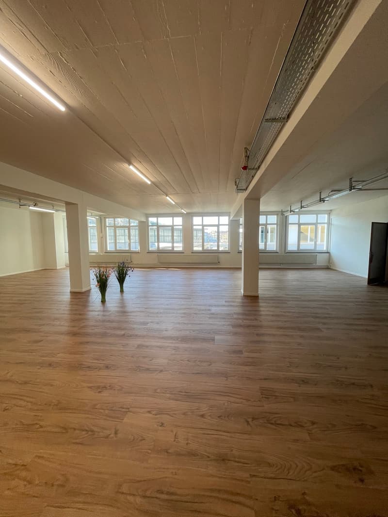 Large room with wooden floors, fluorescent lighting on the ceiling, windows along the perimeter.
