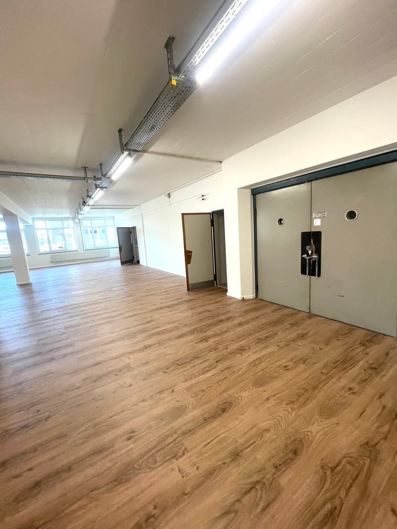 Wide open space with large wooden flooring, fluorescent ceiling lights, three doors, two visible windows