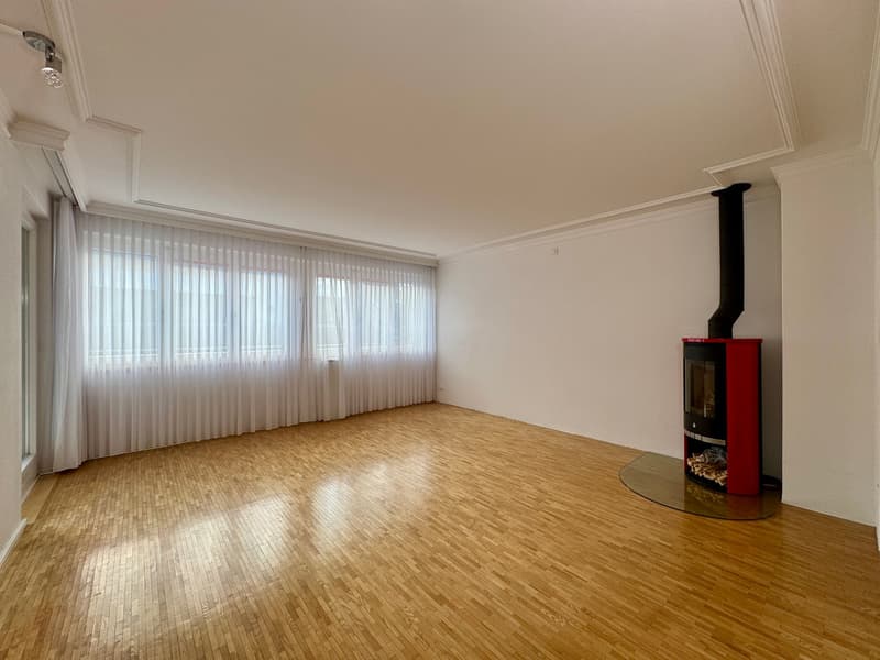 Empty room with hardwood floors, white walls, fireplace in the corner, windows covered with white curtains, spot lights on ceiling