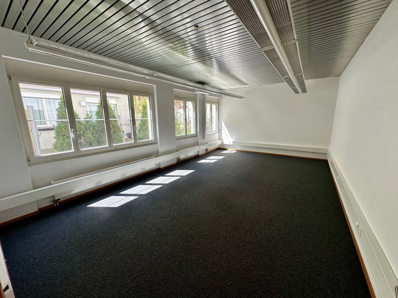 Empty room with white walls, black carpet, large windows, metal ceiling, and white radiators