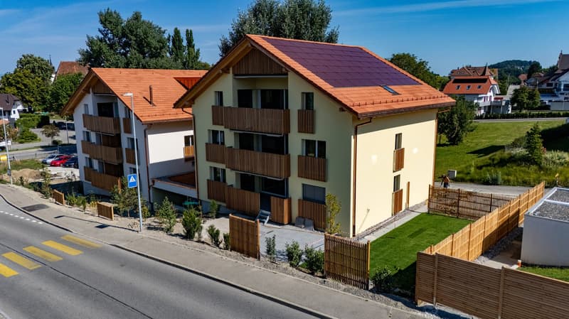 Two-storey house, red roof, yellow exterior, solar panels, wooden balconies, gated entrance, green lawn, road with pedestrian crossing