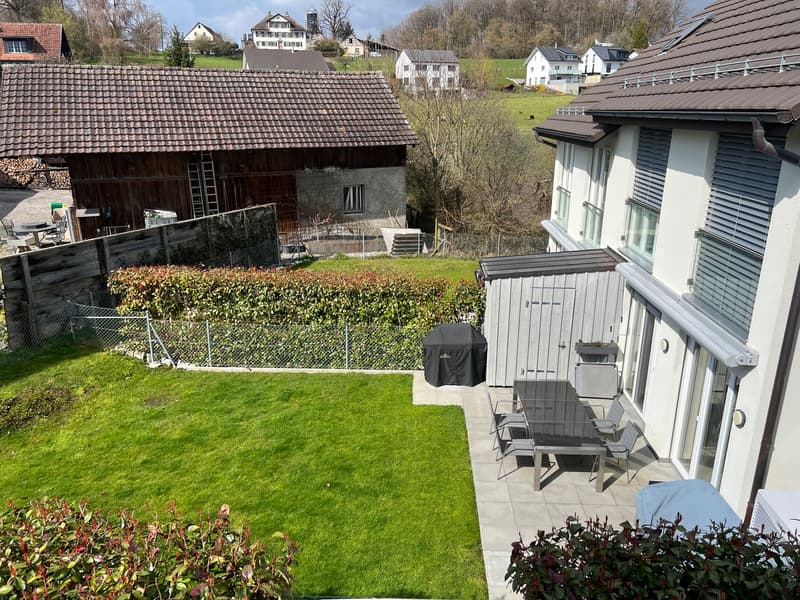 Aerial view of a garden with a table and chairs, a grill cover, and a storage shed. The garden is surrounded by a fence and has a grassy lawn. In the background, there is a house with a brown roof and a barn with a ladder leaning against it. There are als