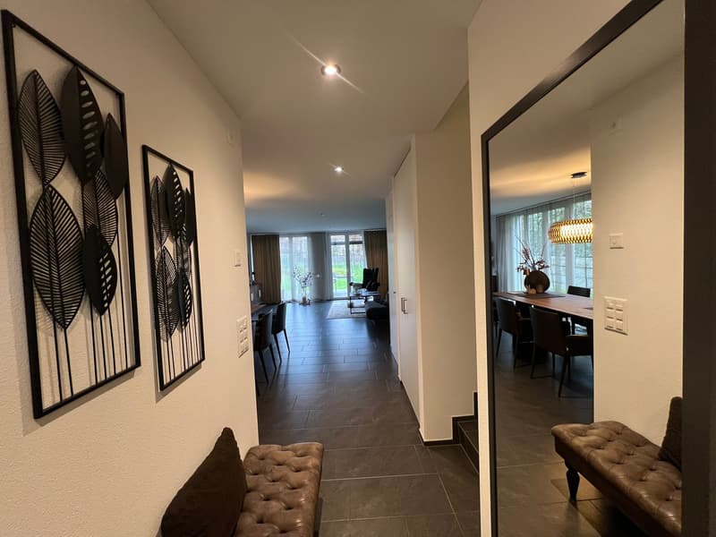A hallway with a bench, two wall art pieces, and a mirror. The hallway leads to a dining area with a long table, chairs, and a chandelier. There is a window with curtains and a view of a garden outside.