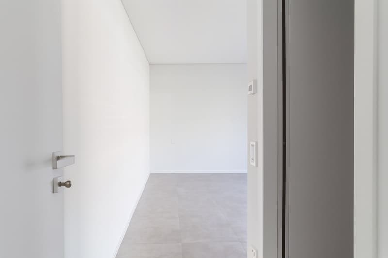 An empty corridor with white walls, grey flooring, and no furniture or objects visible.