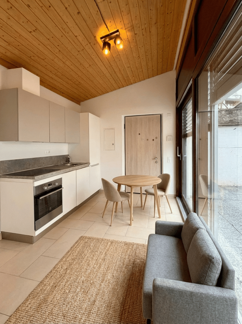 modern open plan kitchen, dining area, and living space with white cabinetry, granite countertops, large windows, wooden dining table, and a gray sofa