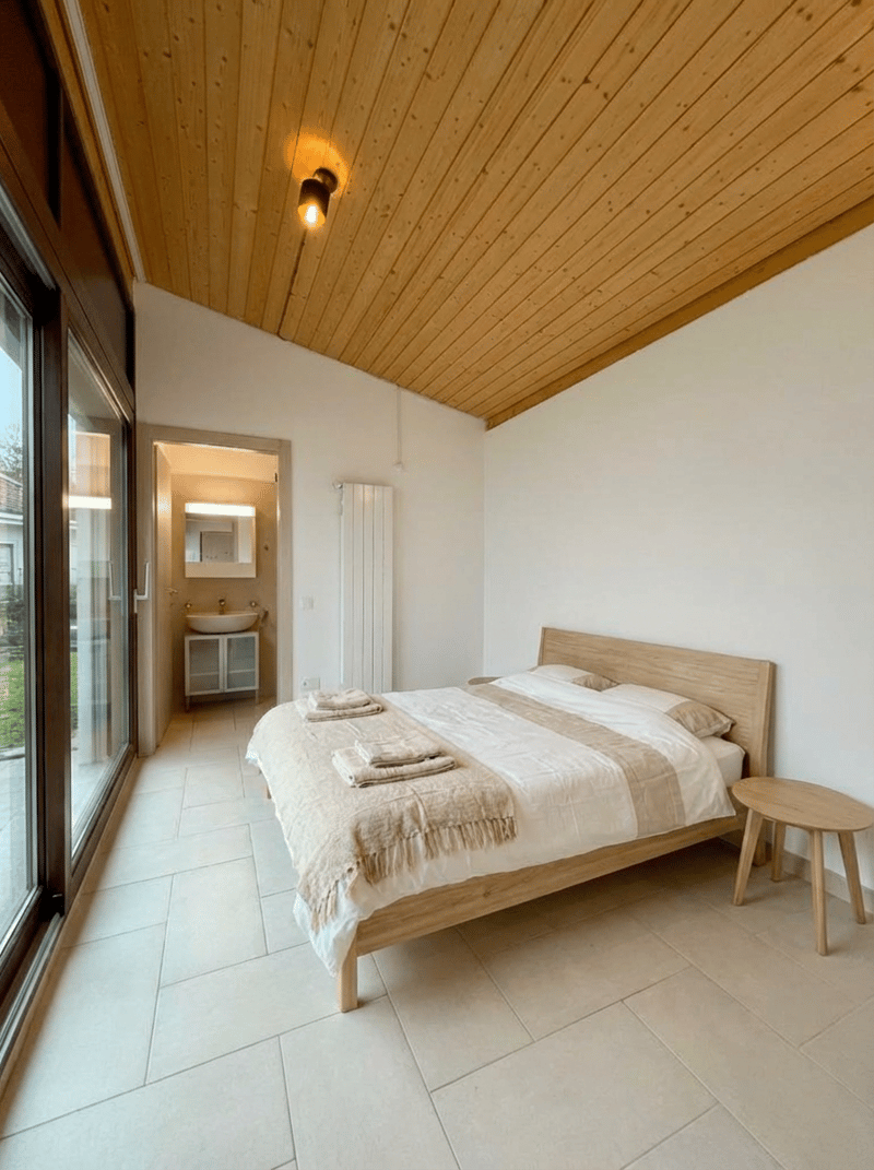 A modern bedroom featuring a double bed with clean, crisp white bedding, wooden side table, wooden ceiling, glass sliding door, adjacent bathroom with sink, small wooden cabinet, and a mirror.