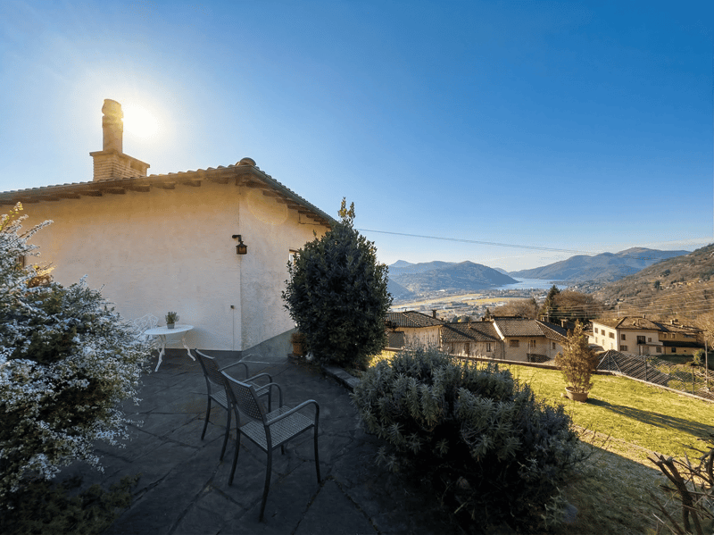 House with a patio, surrounded by plants, a table, and chairs, with a view of the mountains and a lake.