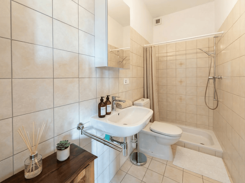 Bathroom with white tiles, white sink, white toilet, shower, wooden cabinet, potted plant, and shower curtain.