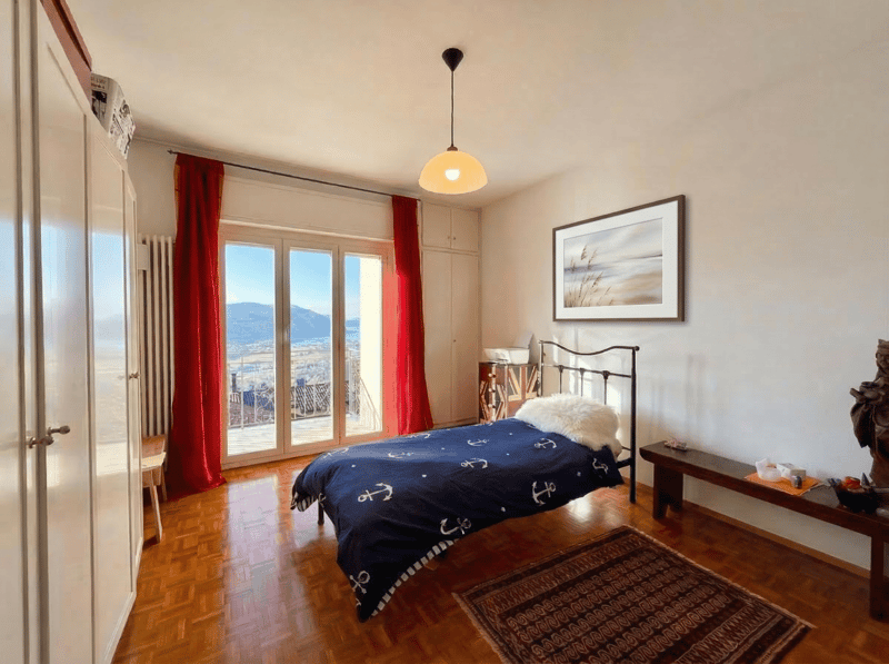 single bed, blue bedspread, wooden floor, large window, sliding glass door, balcony, wooden bench, rug, picture frame, lamp