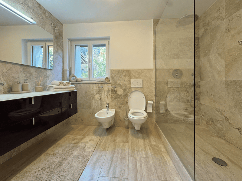 modern bathroom with marble tiles, glass shower enclosure, white toilet, bidet, vanity with mirror, towel rack, and window