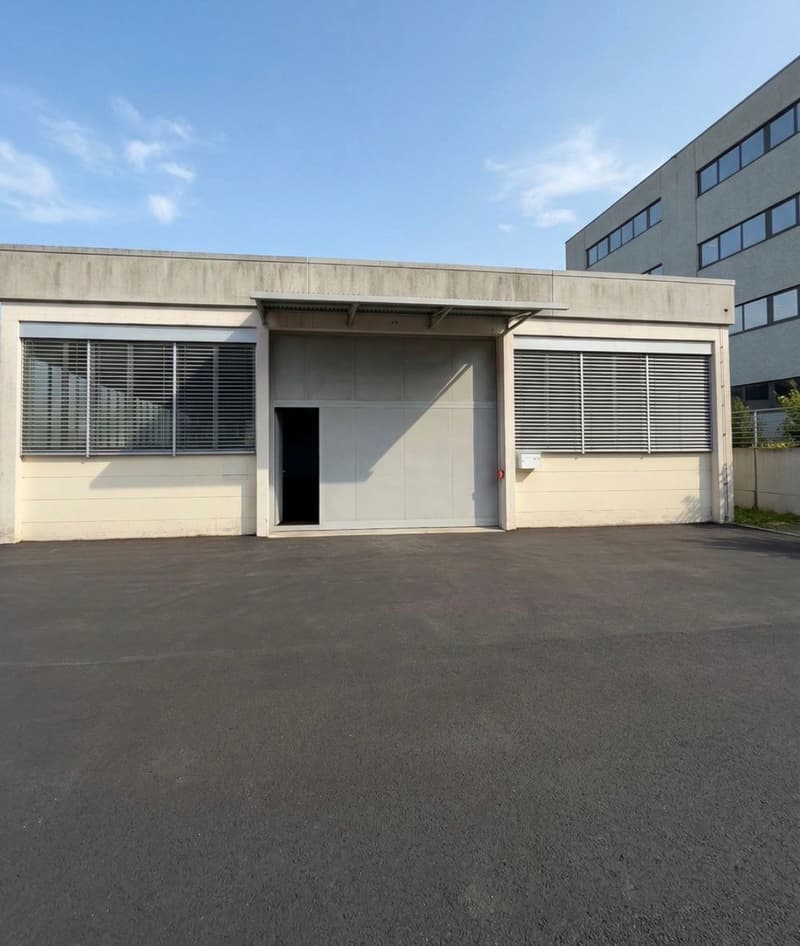 Industrial building with a large garage door, a parking lot in front, and a smaller building in the background.