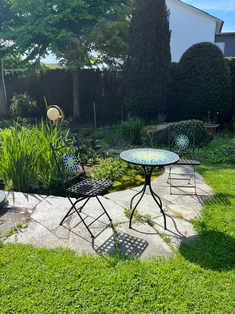 backyard, small round mosaic table, black folding chairs, greenery, trees, stone path, decorative lantern