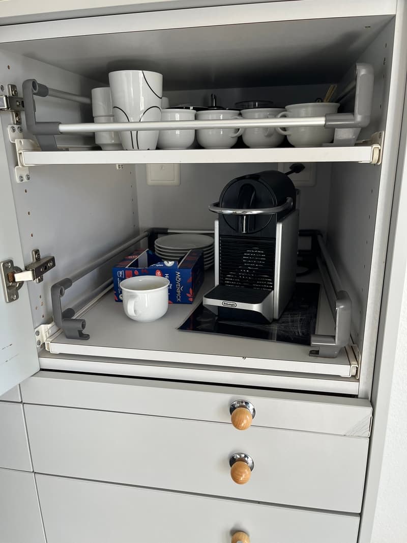 White cabinet with several drawers, stackable cups, plates, coffee machine, box of coffee, and an electrical socket.