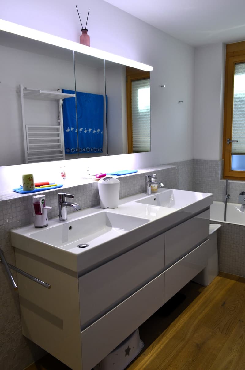 modern bathroom with double sink, bathtub, large mirror, wooden floor, white tiles, towel rack
