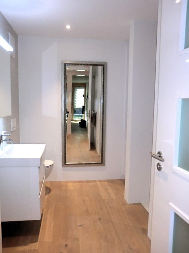 Bathroom with white walls, wooden floor, white sink, mirror, toilet, and a door