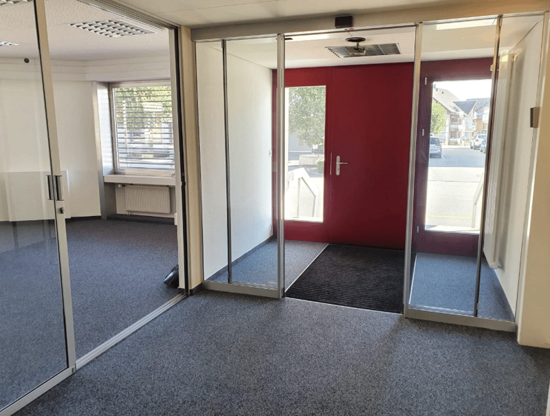An empty room with sliding doors, carpeted floor, a window with blinds, a radiator on the wall, and a mat outside the entrance