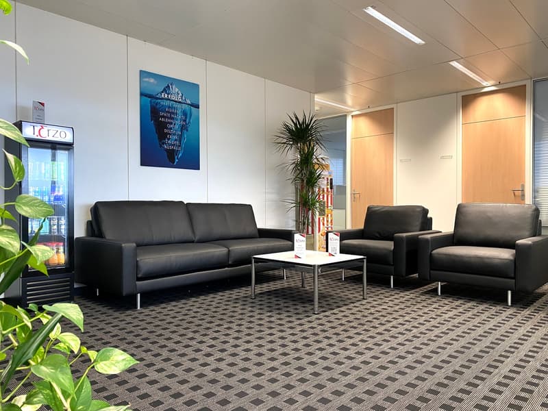 The image shows a modern office lobby with a waiting area. The space features black leather sofas and armchairs arranged around a glass coffee table. The walls are decorated with a large framed poster and a potted plant. The floor is covered with a patter