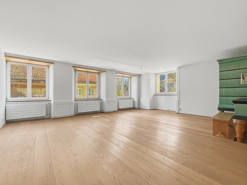An empty room with wooden flooring, large windows, radiators below the windows, a wooden table, and a green vertical storage unit on the right side.