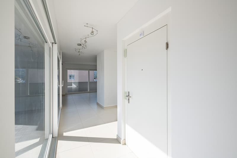 Empty hallway with white tiles, glass doors, and a balcony visible in the distance.
