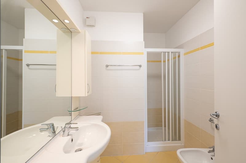 modern bathroom with white tiles, glass shower enclosure, white sink, mirror, and a small cabinet